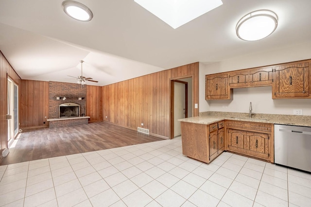 kitchen featuring stainless steel dishwasher, a sink, wooden walls, a ceiling fan, and a fireplace