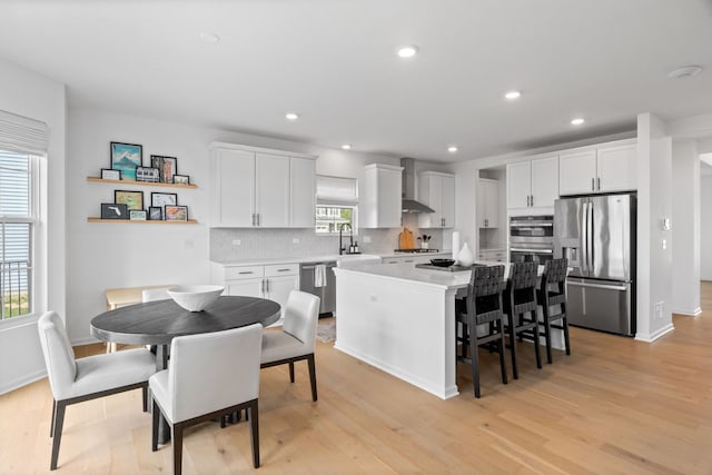 kitchen with stainless steel appliances, wall chimney exhaust hood, a kitchen island, light wood-style floors, and decorative backsplash
