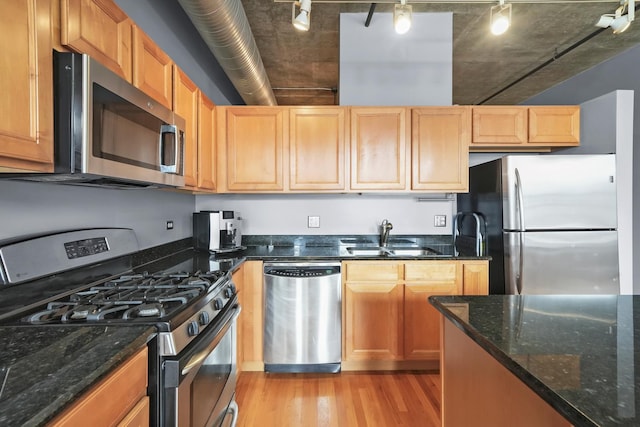 kitchen featuring appliances with stainless steel finishes, a sink, light wood-style floors, rail lighting, and dark stone countertops