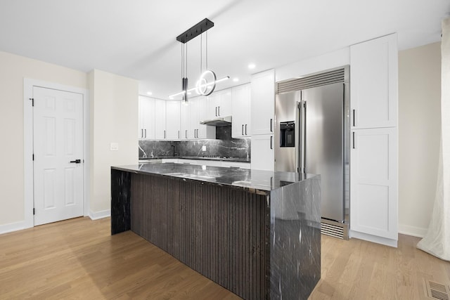 kitchen featuring high end refrigerator, light wood-style floors, a kitchen island, backsplash, and dark stone countertops