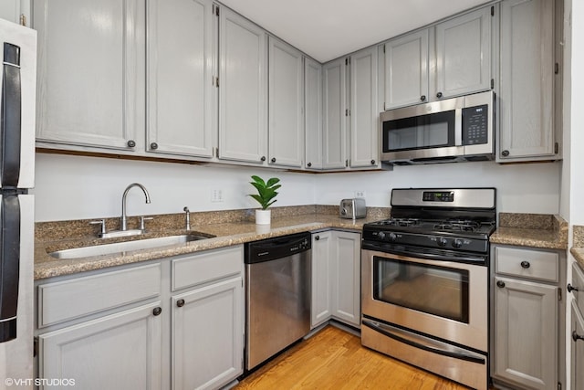 kitchen with stainless steel appliances, a sink, light wood finished floors, gray cabinetry, and stone counters