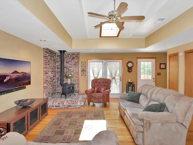 living room featuring a wood stove, ceiling fan, light wood-style flooring, and a raised ceiling
