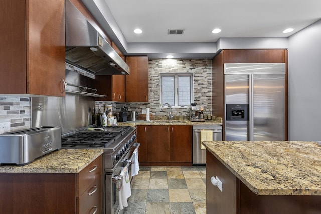 kitchen featuring wall chimney range hood, high quality appliances, light stone finish flooring, decorative backsplash, and light stone countertops