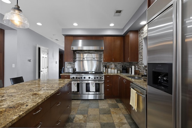 kitchen featuring appliances with stainless steel finishes, a sink, wall chimney exhaust hood, stone finish flooring, and backsplash