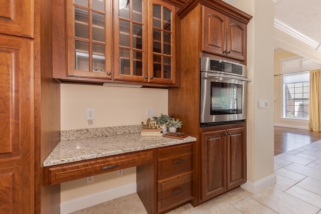 kitchen featuring stainless steel oven, light stone counters, glass insert cabinets, light tile patterned floors, and brown cabinetry