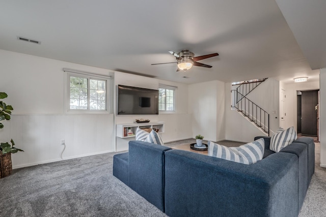 living room with carpet flooring, stairs, a ceiling fan, and a wainscoted wall