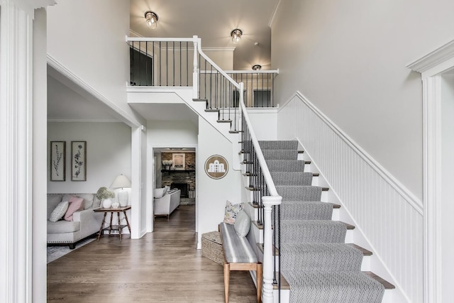 staircase with a high ceiling, a stone fireplace, crown molding, and wood finished floors