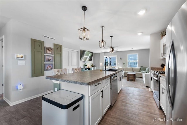 kitchen with stainless steel appliances, a sink, open floor plan, dark countertops, and a center island with sink
