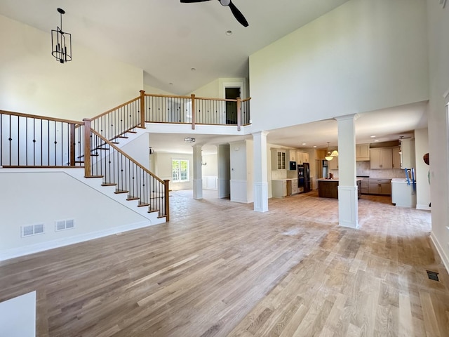 unfurnished living room with decorative columns, ceiling fan, a high ceiling, light wood-style floors, and stairway