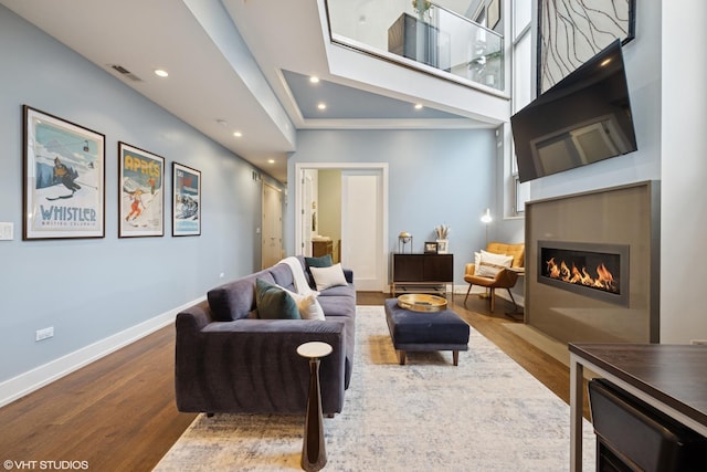 living area with wood finished floors, a glass covered fireplace, baseboards, recessed lighting, and a high ceiling