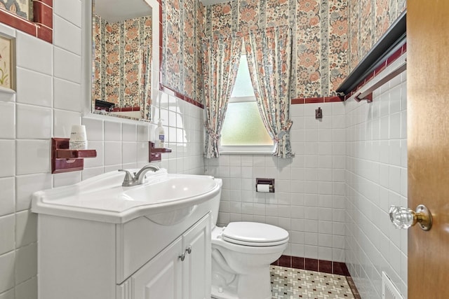 bathroom with tile walls, toilet, vanity, a wainscoted wall, and wallpapered walls