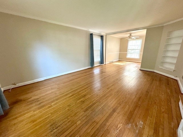 empty room with built in shelves, crown molding, wood finished floors, ceiling fan, and baseboards