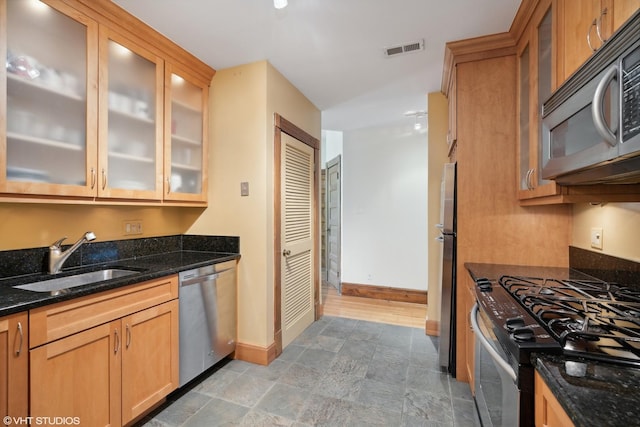 kitchen with appliances with stainless steel finishes, a sink, baseboards, glass insert cabinets, and dark stone countertops
