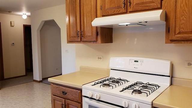 kitchen featuring white gas range, under cabinet range hood, brown cabinetry, light countertops, and arched walkways