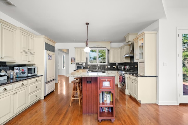 kitchen featuring high end appliances, cream cabinets, wall chimney exhaust hood, and a kitchen bar