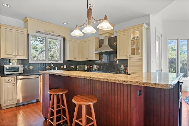 kitchen with cream cabinets, stainless steel dishwasher, wall chimney exhaust hood, and a kitchen bar