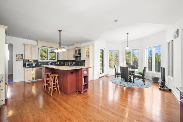 kitchen with cream cabinetry, wall chimney exhaust hood, wood finished floors, stainless steel dishwasher, and backsplash