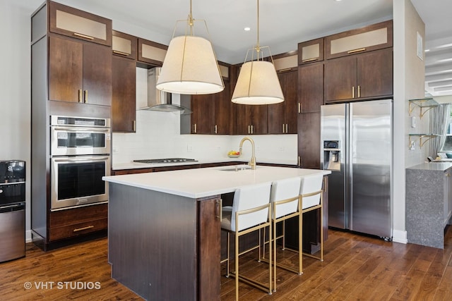 kitchen featuring appliances with stainless steel finishes, wall chimney range hood, dark wood finished floors, decorative backsplash, and a center island with sink