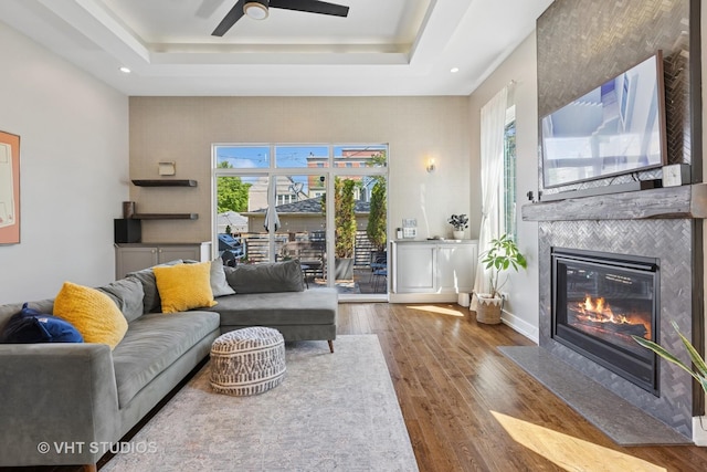 living room with ceiling fan, a raised ceiling, wood finished floors, a large fireplace, and recessed lighting