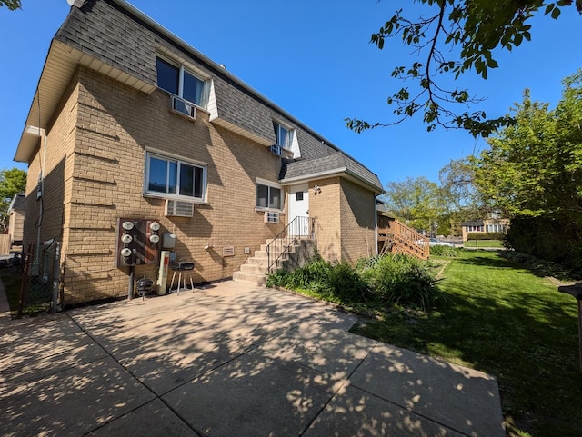 back of house with mansard roof, roof with shingles, brick siding, a lawn, and a patio