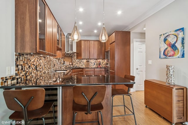 kitchen featuring wall chimney range hood, a peninsula, decorative backsplash, glass insert cabinets, and brown cabinets