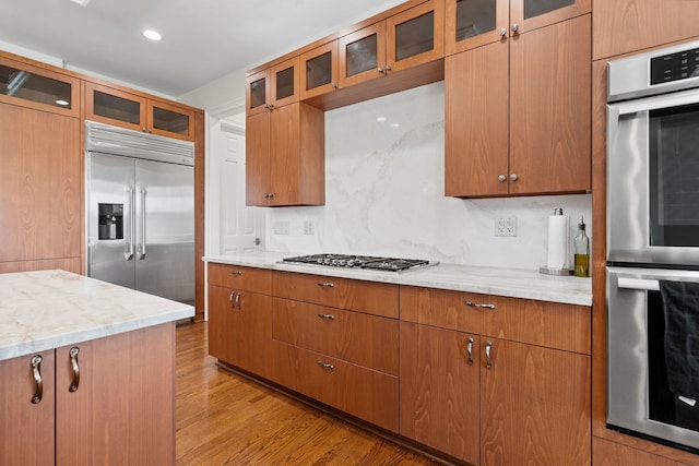 6590 Beech Ln, Libertyville, IL 60048 - kitchen featuring stainless steel appliances, light wood-type flooring, brown cabinets, decorative backsplash, and glass insert cabinets
