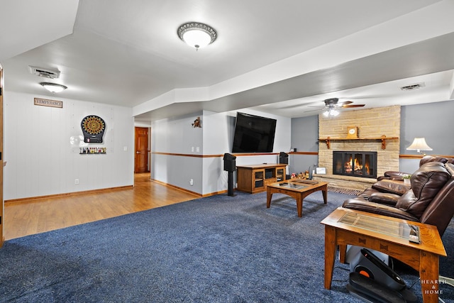 living room with baseboards, wood finished floors, a ceiling fan, a stone fireplace, and carpet flooring