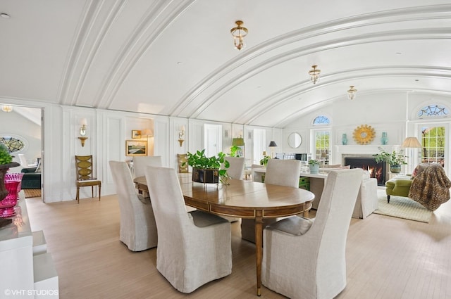 dining area featuring healthy amount of natural light, light wood-type flooring, a warm lit fireplace, and lofted ceiling