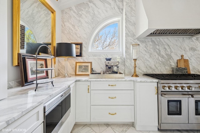 kitchen with appliances with stainless steel finishes, wall chimney exhaust hood, white cabinets, light stone countertops, and backsplash