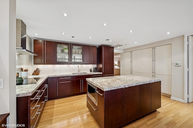 kitchen with a sink, wall chimney range hood, stainless steel microwave, light wood-style flooring, and tasteful backsplash