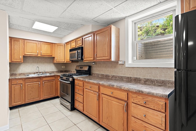 kitchen with appliances with stainless steel finishes, a sink, a drop ceiling, light tile patterned floors, and dark countertops