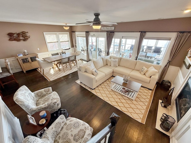 living room with a ceiling fan, healthy amount of natural light, dark wood finished floors, and wainscoting