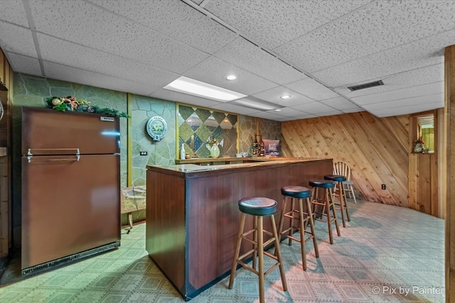 indoor bar featuring freestanding refrigerator, tile patterned floors, a drop ceiling, and wood walls
