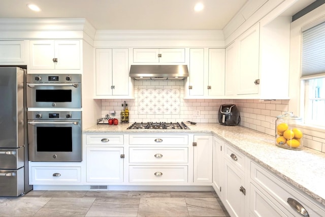 kitchen featuring appliances with stainless steel finishes, under cabinet range hood, white cabinets, light stone counters, and backsplash