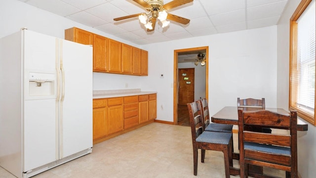 kitchen featuring white fridge with ice dispenser, a ceiling fan, light countertops, a drop ceiling, and brown cabinets