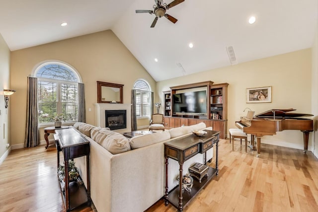 living area featuring high vaulted ceiling, ceiling fan, light wood finished floors, a glass covered fireplace, and baseboards