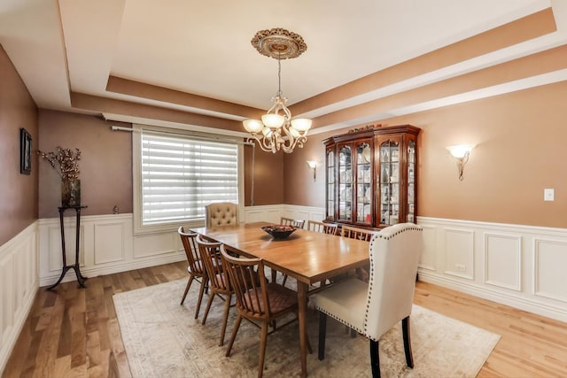 dining room with a chandelier, light wood finished floors, wainscoting, a raised ceiling, and a decorative wall