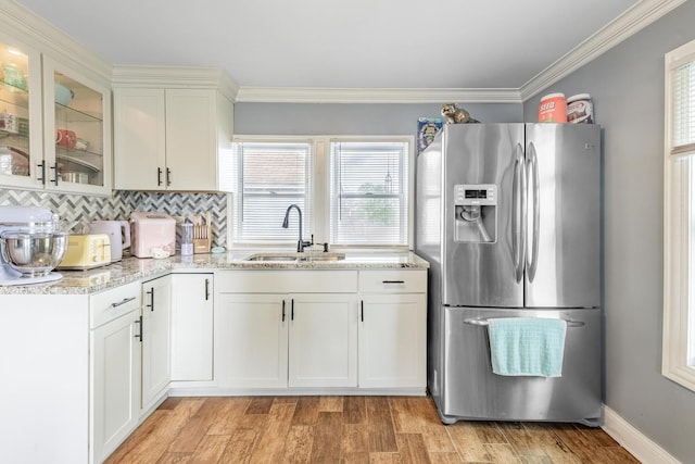 kitchen with stainless steel refrigerator with ice dispenser, a sink, glass insert cabinets, light stone countertops, and crown molding