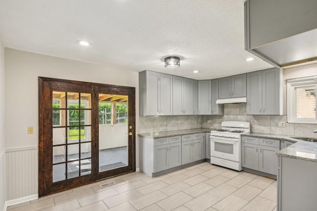kitchen with white gas range oven, gray cabinetry, healthy amount of natural light, light stone counters, and wainscoting