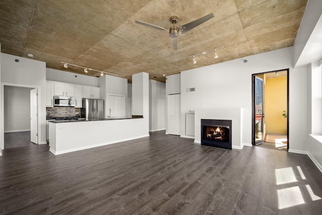 unfurnished living room with rail lighting, dark wood-style floors, ceiling fan, a lit fireplace, and baseboards