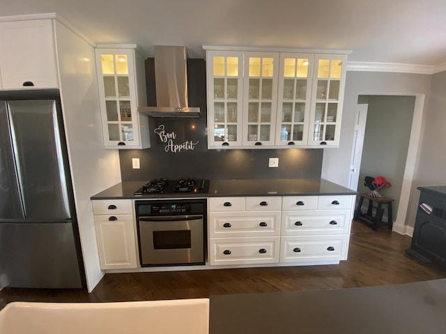 kitchen with appliances with stainless steel finishes, wall chimney exhaust hood, dark countertops, ornamental molding, and white cabinetry