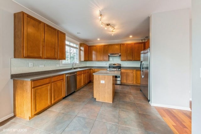 kitchen with appliances with stainless steel finishes, a sink, under cabinet range hood, a kitchen island, and brown cabinetry