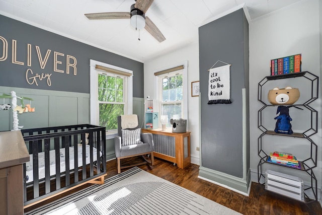 bedroom with wood finished floors, crown molding, a crib, a wainscoted wall, and a ceiling fan