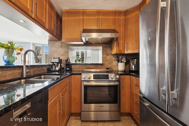 kitchen with stainless steel appliances, a sink, under cabinet range hood, backsplash, and dark stone countertops
