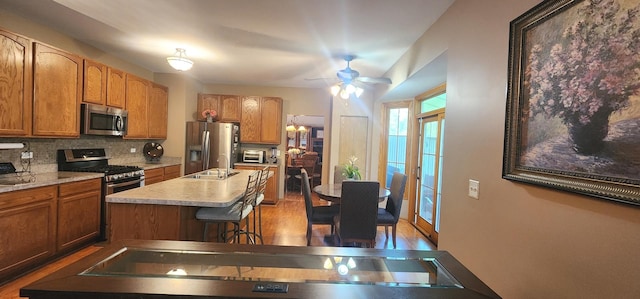 kitchen featuring stainless steel appliances, a sink, decorative backsplash, a center island with sink, and brown cabinets