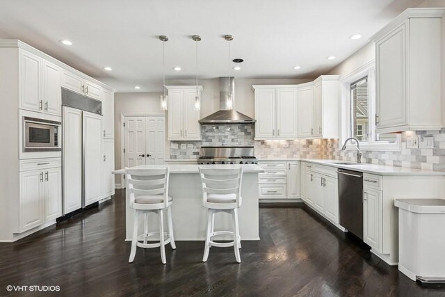 kitchen featuring wall chimney range hood, built in appliances, a sink, a kitchen island, and tasteful backsplash