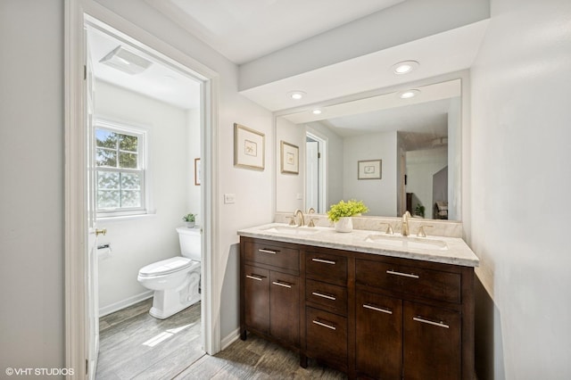 bathroom featuring double vanity, toilet, wood finished floors, baseboards, and recessed lighting
