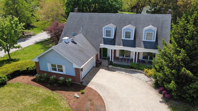 new england style home featuring covered porch, decorative driveway, brick siding, a garage, and a shingled roof