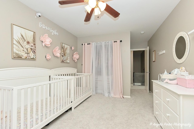 bedroom with a crib, light colored carpet, a ceiling fan, and baseboards
