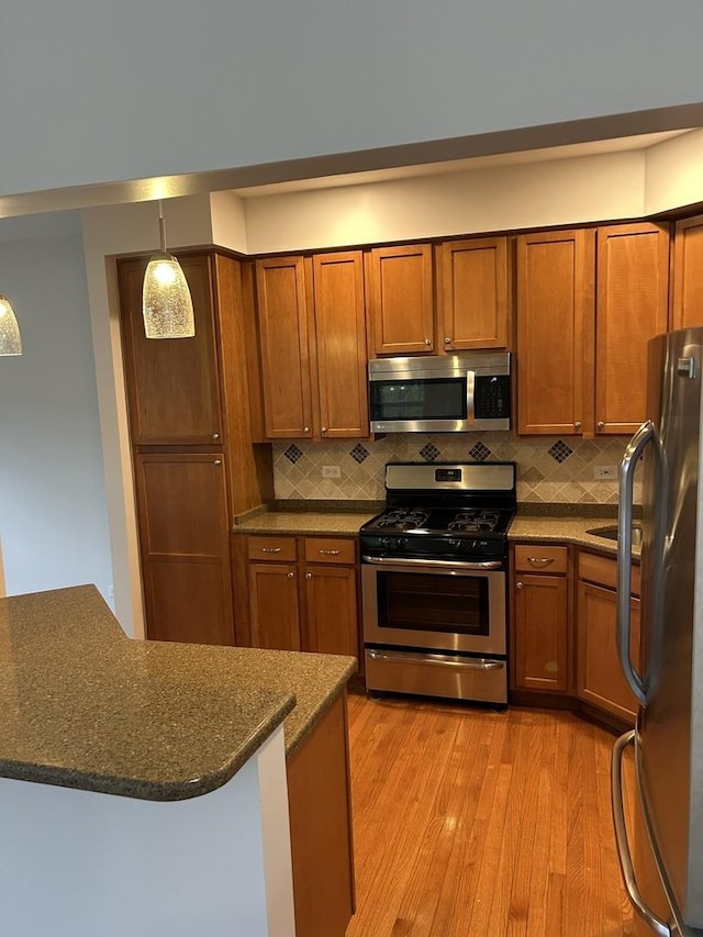 kitchen with appliances with stainless steel finishes, brown cabinetry, decorative backsplash, light wood-type flooring, and dark stone countertops